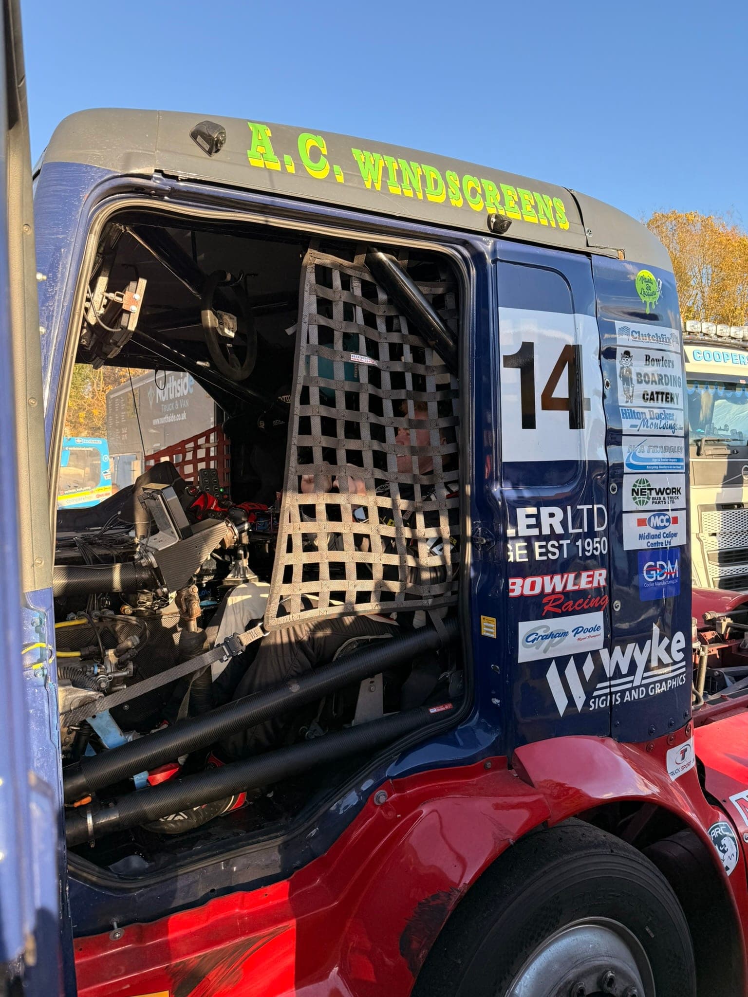 Diana’s open cab in bright sun, the window net fastened and the driver strapped in behind it, a column of partner logos on the door