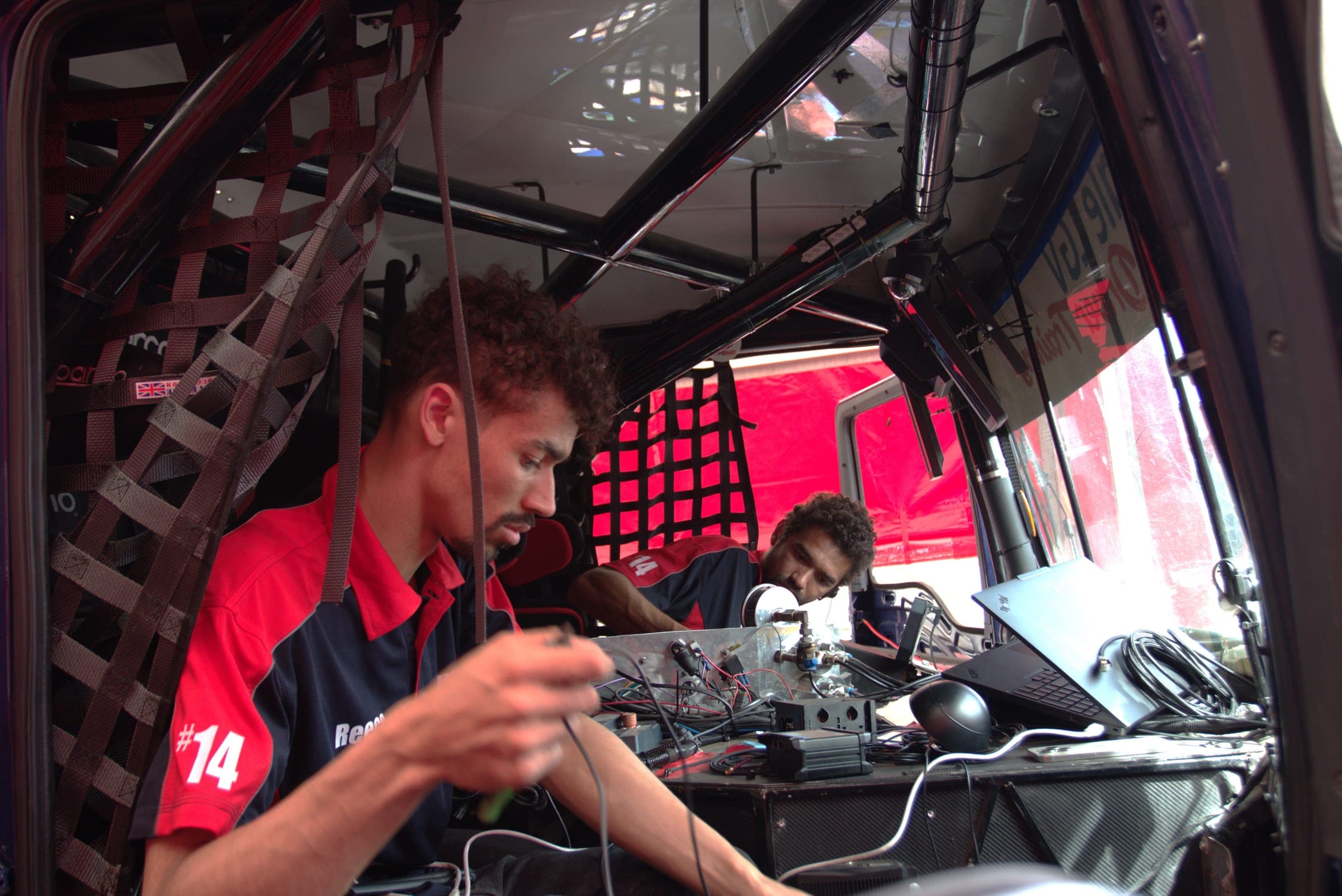 Two crew members working on wiring and a laptop inside Diana’s stripped race cab, surrounded by the roll cage
