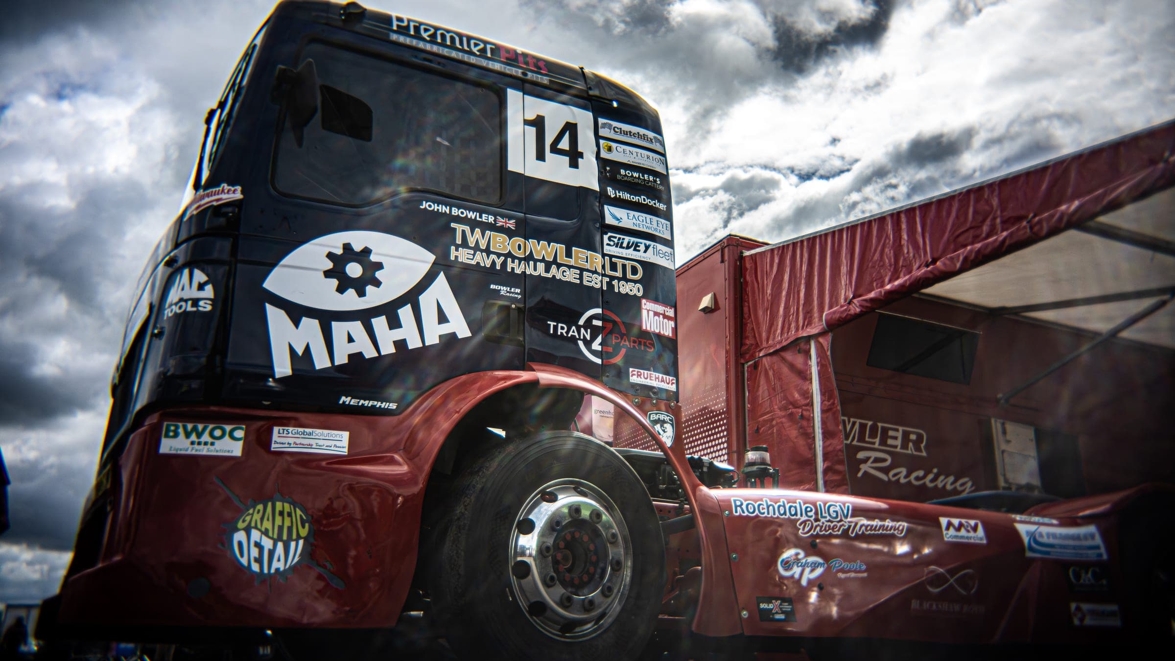 Low view of Diana’s cab beside the red Bowler Racing awning under dramatic cloud, John Bowler’s name on the door