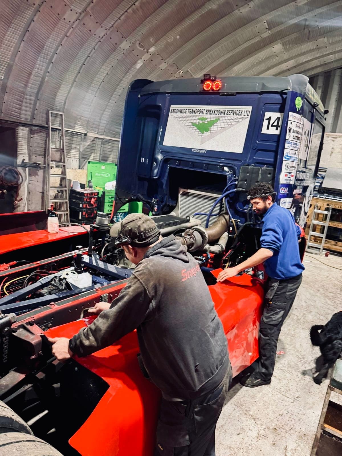 Two crew members fitting red bodywork to Diana in an arched workshop, a black dog at their feet