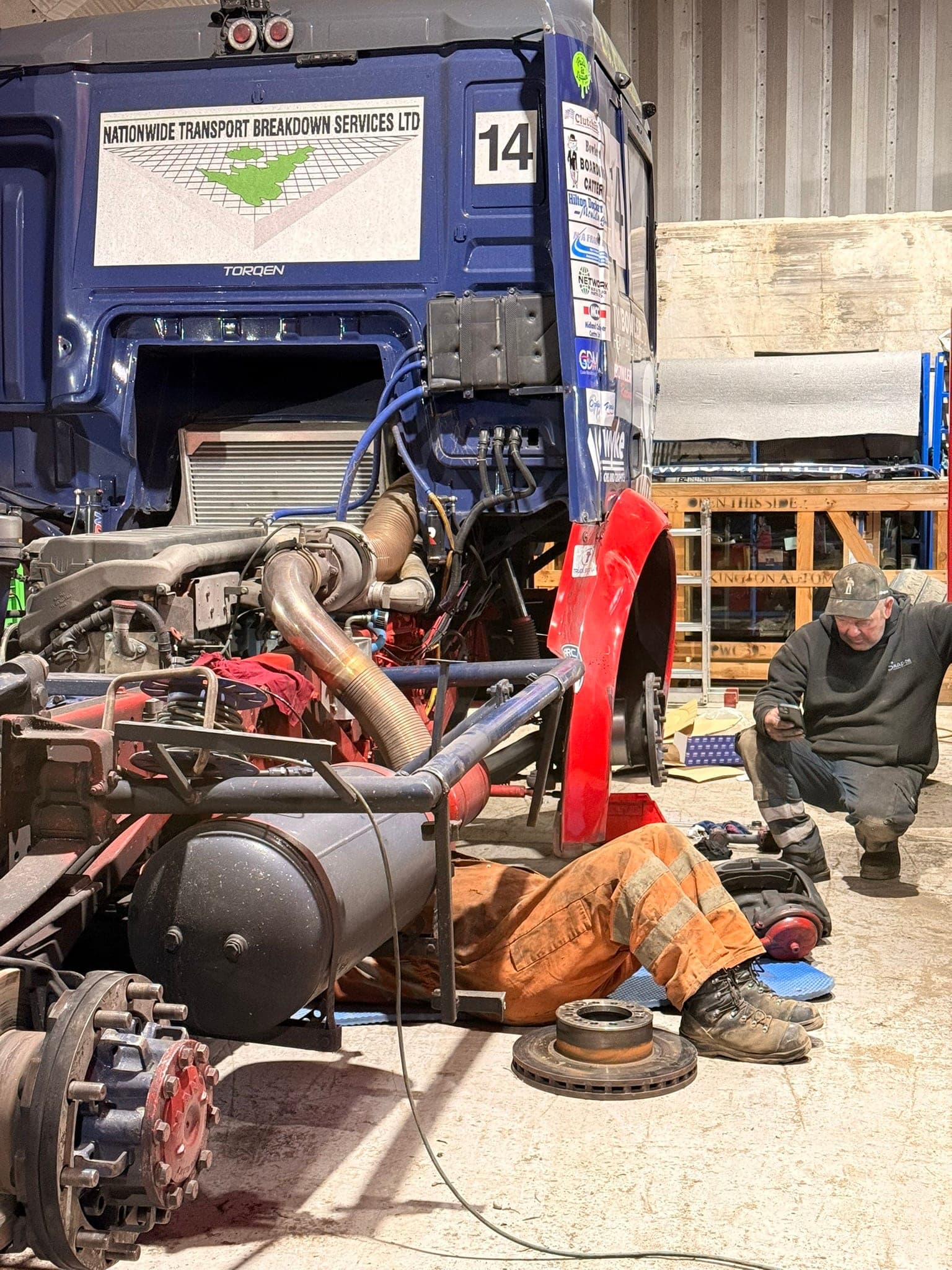 Diana stripped in the workshop, a mechanic in orange overalls underneath and another crouched checking a phone